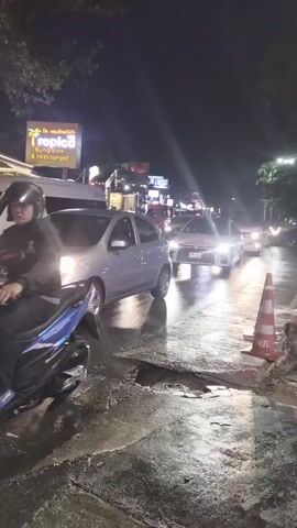 The media shows a nighttime scene on a wet street in Patong, Thailand. A silver car is in the foreground, with a motorcycle approaching from the background. Several other vehicles, including cars and motorcycles, are visible on the road, some with their headlights on, illuminating the wet asphalt. The street appears to be in disrepair, with a large pothole visible in the road. Orange traffic cones are placed on the sidewalk, indicating a work area or hazard.

In the background, there are buildings with illuminated signs, suggesting a commercial or entertainment district. One sign visible reads "TACO BANGALOW RESTAURANT." The lighting suggests it is nighttime, and the wetness of the road indicates recent rain or damp conditions. The overall atmosphere is that of a busy, urban street at night, with traffic and pedestrian activity, despite the poor road conditions. The scene captures a common urban environment with an element of everyday challenges like road maintenance.