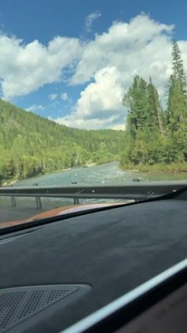 The video shows a view from inside a car driving along a scenic route next to a wide river. The river flows through a valley surrounded by lush green, tree-covered mountains. The sky is blue with scattered white clouds. On the right side of the road, a white traffic sign with a red border displays a symbol of falling rocks and the text "1500m". The camera is positioned to show the dashboard and the windshield of the car. The sunlight appears to be bright, indicating a clear day. The car is moving, as suggested by the slight blur and the changing perspective of the scenery. The road is bordered by a metal guardrail. The overall impression is one of a peaceful and beautiful drive through a natural landscape.