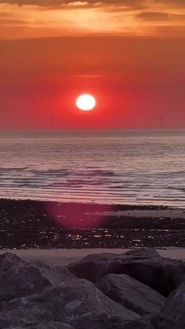 A vertical video shows a vibrant sunset over the ocean. The sun is a bright, white orb positioned in the center of the frame, casting a powerful red glow across the sky. The sky transitions from a deep red near the horizon to a warm orange and yellow at the top. Gentle waves roll onto a dark, pebbly shore in the foreground. Large, dark rocks form the very bottom of the frame. In the distance, silhouetted against the horizon, are several wind turbines, suggesting an offshore wind farm. The water reflects the colorful hues of the sunset, creating shimmering patterns. The scene is peaceful and serene, capturing the dramatic beauty of the end of the day.