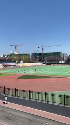 The video shows an outdoor athletic field with a red running track surrounding a green field. Two people are on the field near a soccer goal. One person is wearing a red shirt and the other is wearing a white shirt. Several soccer balls are scattered on the field. In the background, construction is taking place with multiple cranes visible against a clear blue sky.  A black fence runs along the bottom of the frame, separating the field from a sidewalk. Two people are walking along the sidewalk. The day appears bright and sunny.