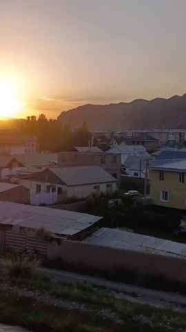 This video captures a panoramic view of a residential area in Naryn City, Kyrgyzstan, during sunset. The scene is dominated by a cluster of houses with varied architectural styles, many featuring sloped roofs made of corrugated metal or tiles. A prominent yellow two-story building with white-framed windows stands out in the foreground, suggesting a modern addition to the otherwise more traditional housing. Beyond these, a collection of apartment buildings with multiple stories are visible, indicating a more urbanized section of the city. In the distance, a majestic mountain range forms the backdrop, its slopes bathed in the warm, fading light of the setting sun. The sky transitions from a soft yellow near the horizon to a pale orange and then a dusky blue, characteristic of twilight. A golden-domed building, likely a mosque, is visible in the middle ground, its illuminated dome catching the last rays of sunlight, adding a spiritual element to the landscape. The overall atmosphere is serene and peaceful, with the natural beauty of the mountains contrasting with the human settlement. The time of day is clearly indicated by the low angle of the sun and the soft, diffused light. There is no visible text in the video. The setting appears to be a quiet evening in a semi-urban environment nestled amongst a vast natural landscape.