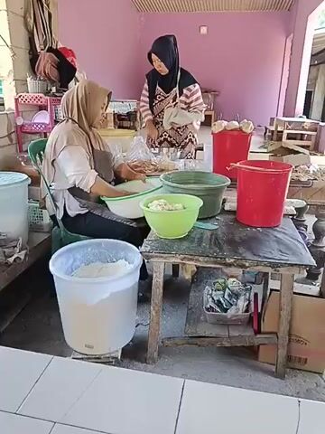 The video shows two women in a room, likely a kitchen or food preparation area. The woman in the foreground, wearing a light brown headscarf and apron, is seated on a chair. She is mixing something in a large white bowl. In front of her are several bowls of ingredients, and a large white plastic container filled with a white powdery substance. To her left is a large light blue container. The woman in the background, wearing a dark hijab and an apron with a checkered pattern, is standing and appears to be chopping or preparing food on a table. There are red buckets and other containers on the table, as well as bags of food items and some white spherical objects. The room has pink walls and a wooden ceiling with some sort of screening material. There are shelves in the background with various items. The overall impression is of a food production or cooking environment.