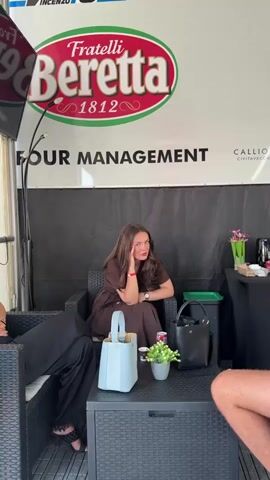 The video begins with a wide shot of a woman sitting in a chair outdoors, possibly at an event or venue, with a large banner behind her that says "Fratelli Beretta 1812" and "TOUR MANAGEMENT". She is wearing a brown dress and has her hand near her face. The shot then zooms in on her face, where she is looking at the camera with a serious expression. Her hand is touching her face, and she appears to be making a gesture or pose. The background is a dark curtain. There are other objects visible in the wider shot, including a light blue purse, a black purse, a small plant, and a drink can on a table in front of the woman. The lighting is bright, and the focus is on the woman.