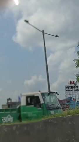The video captures a daytime view from a moving vehicle, likely a car or bus, on a highway. The scene is an urban landscape with buildings, trees, and flags visible. The weather appears to be partly cloudy with bright sunlight.

The subjects are primarily buildings of various colors, including a prominent blue building with a pointed, green-topped structure. Several flags are displayed, mostly white with blue stripes, and some are green. The flags are attached to poles and are seen on top of buildings, suggesting a celebration or political event.

The activity is the movement of the vehicle through the area, indicated by the shifting perspective and the blurred foreground of vegetation and a guardrail. The time of day is clearly daytime due to the bright sunlight and shadows. The location is Aminbazar, Bangladesh, as indicated by the provided context.

Notable details include the presence of a lamppost on the right side of the frame, and dense green foliage in the foreground. The sky is a bright blue with fluffy white clouds. There is no visible text in the frame. The overall mood is neutral, documenting a passing scene.