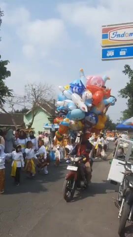 A parade is in progress on a paved road under a bright, partly cloudy sky. In the foreground, a person rides a red motorcycle, carrying a large bunch of colorful character balloons. The balloons are attached to the motorcycle's handlebars and are a mix of shapes and characters, including Doraemon, a pink creature, a red fish-like character, and a blue bear. The parade participants, many of whom are children wearing traditional white and gold clothing with patterned sashes, are walking down the road. Some adults are also present, some in similar attire and others in more casual clothing. To the right, a white sign with an "Indomaret" logo is visible. The environment appears to be a rural or suburban area with houses and trees lining the road. The overall atmosphere seems festive.