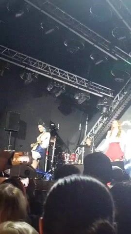 A short video shows a live music performance. The stage is lit with green lights. A woman in a white top and blue shorts is singing and dancing. She is wearing knee-high blue boots. A man in a black shirt is standing to her left. A drum set is visible behind her. The audience is in the foreground, with some people holding up phones to record the performance.