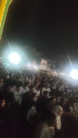 The video shows a large crowd of people gathered outdoors at night. The scene is illuminated by bright spotlights, creating a bright, hazy effect. The crowd appears to be a mix of men and women, with many of them wearing traditional Islamic clothing. The people are facing in the same direction, suggesting they are watching a performance or listening to a speaker. The atmosphere appears to be one of reverence or celebration.