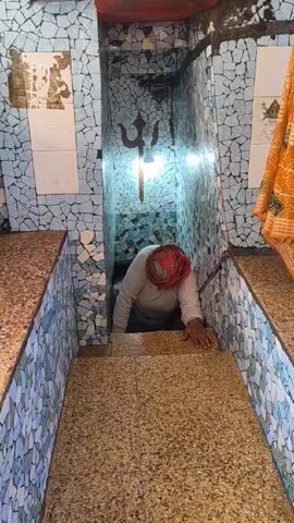A woman with a red headscarf is seen descending a narrow staircase. She is wearing a white shirt and using her hands for support as she navigates the steps. The staircase is flanked by tiled walls with a mosaic pattern in shades of blue and white. On one wall, a trident symbol is visible above a light fixture. The lighting is moderate, and the space appears to be enclosed. There is a patterned cloth draped on the right side of the frame.