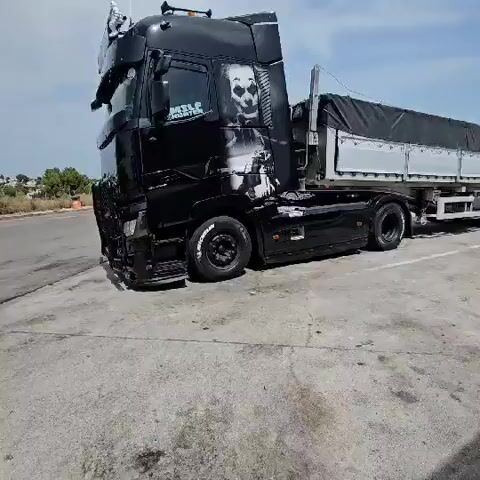 The video shows a black semi-truck parked in an outdoor lot. The truck is black with a graphic design on the side, including a panda face. The truck has a large grill guard in the front. Other trucks are visible in the background. The ground is paved with concrete and there is some debris present. The sky is visible in the background, with some clouds. The camera appears to be handheld and the footage is slightly shaky.