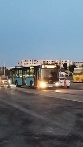 A blue bus with its headlights on is parked on a paved road. The bus has its doors open. Orange and white traffic cones are placed on the road. Several cars are also visible on the road, some with their headlights on. Buildings are in the background. The sky is a dusky blue.