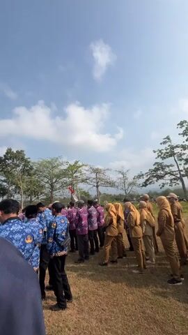 A crowd of people, many wearing matching blue and purple patterned shirts and khaki uniforms, stand outdoors under a bright blue sky with scattered clouds. The people are mostly facing away from the camera, suggesting a formal gathering or event. Some individuals are wearing headscarves. In the background, there are trees and a red and white flag on a pole. The ground appears to be dry grass or dirt. The overall atmosphere seems to be one of an official assembly.
