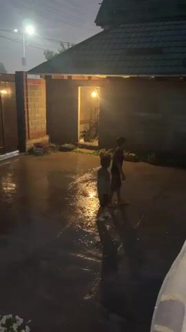 This video captures a nighttime scene in what appears to be a backyard or courtyard in Komsomolskoe, Kyrgyzstan. Two young children are visible, one in a dark t-shirt and shorts, the other in a camouflage t-shirt and dark shorts. The ground is wet, indicating recent rain, and puddles reflect the warm glow of a single light source emanating from an open doorway of a brick structure. This structure, possibly a shed or garage, has a dark, tiled roof and is dimly lit from within. A streetlamp casts a cooler light in the distance, illuminating a segment of a brick wall and a fence. The sky is dark, suggesting it is either late evening or early morning. The overall mood is tranquil and slightly mysterious, with the children engaged in playful movement on the wet ground. There is no visible text in the video.
