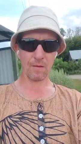 A man wearing a bucket hat, sunglasses, and a brown t-shirt with a black print is filmed while riding a bicycle. The video also shows a rural setting with houses and trees in the background. The man is looking directly at the camera and appears to be speaking. The footage is shot from a first-person perspective, indicating it was likely taken using a phone or action camera.