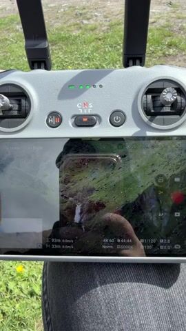 The video shows a DJI drone remote controller held in front of a person wearing blue jeans. The drone is in flight, and the controller screen displays a live view of a waterfall in a green, mountainous area. The time is around 19:20. The drone is recording in 4K at 60 frames per second. The area surrounding the person holding the controller has grass and dandelions.