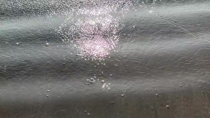 A close-up, high-angle shot focuses on the surface of dark, wet sand as a gentle wave washes over it, reflecting the bright sunlight. The water creates shimmering patterns on the sand, and a thin line of foam forms and recedes with the wave. The camera is stationary.