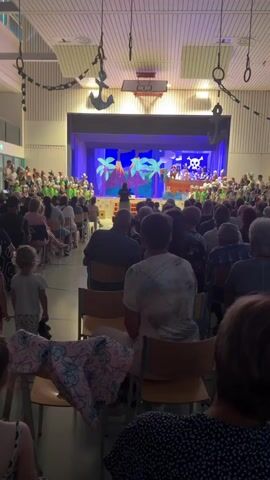 The video shows an indoor event with an audience seated on chairs facing a stage. The stage is decorated with a tropical theme, featuring palm trees, a volcano, and a pirate skull and crossbones. A group of performers, possibly children, are on the stage, some wearing green outfits. Some members of the audience are visible from behind, facing away from the camera. The environment appears to be a hall or gymnasium, with chains and anchors hanging from the ceiling as decorations, suggesting a pirate theme. The lighting is dim, with spotlights on the stage. There is a single person on stage near the front, possibly a speaker or director. The overall atmosphere seems to be that of a performance or school play.