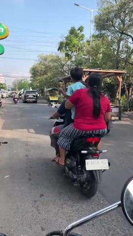 A street scene with multiple vehicles and pedestrians. In the foreground, a person is riding a scooter, facing away from the camera. To the left, another scooter carries two people. Ahead, a three-wheeled vehicle with a canopy is driving. Further down the road, other vehicles and pedestrians are visible. The environment appears to be a sunny day on a paved road with trees and buildings lining the sides.