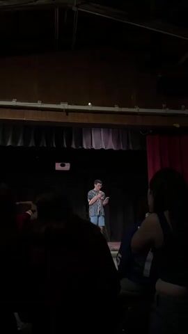 A young person stands on a stage in a dimly lit room, holding a microphone and a phone, and reading from it. The person is wearing a striped shirt and shorts. There are dark curtains behind them, and a projector is mounted on the ceiling. In the foreground, several people are seated in the dark, with their backs to the camera. One person in the foreground is holding a red cup. The room appears to be some kind of auditorium or performance space.