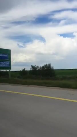 The video displays a landscape scene captured from a moving vehicle, likely a car, on a paved road with a yellow line marking the lane. The time of day appears to be daytime, indicated by the bright sky. The weather seems to be partly cloudy, with a mix of white and gray clouds against a blue sky. The setting is rural, featuring vast green fields that stretch towards the horizon. Bushes and trees are visible along the edge of the road and in the distance. The overall mood is one of travel and open spaces. No people or specific objects are discernible beyond the road and the natural landscape. The location is identified as Osakarovka, Kazakhstan. No text is visible in the video.