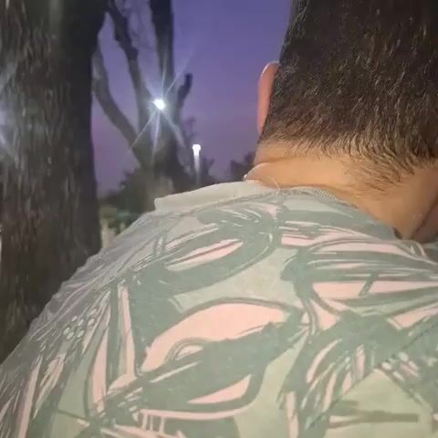 A short video shows a man's head from behind, then his face turning towards the camera. He has short, dark hair and is wearing a light green t-shirt with a leafy pattern. He looks surprised, then smiles broadly, showing his teeth. The background is dark, with trees and lights visible, suggesting it is evening or night.
