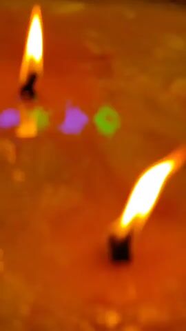 The video shows a close-up view of two lit candles burning in what appears to be melted wax. The wax is a vibrant yellow-orange color, and the surface has a textured, somewhat uneven appearance. The flames of the candles are bright and wavering, casting reflections on the wax surface. The reflections show hints of purple and green light, possibly from ambient lighting or the wax itself. The wicks of the candles are dark and appear to be burning down. The camera angle is a top-down perspective, focusing on the flames and the wax. The video is short, with each frame showing only a slight change in the flames and reflections.