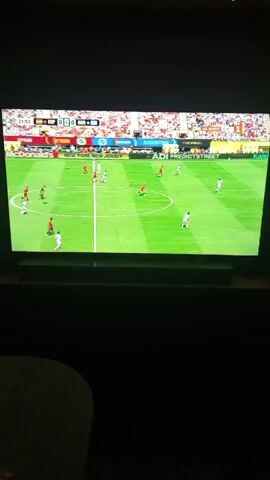 A widescreen television displays a soccer match. The scoreboard at the top of the screen reads "ESP 0 - 0 ARG 29:53". There are multiple players on the green grass field wearing red and white uniforms. The crowd in the stadium is visible in the background. The television screen is framed by a dark area, suggesting it is in a dimly lit room.