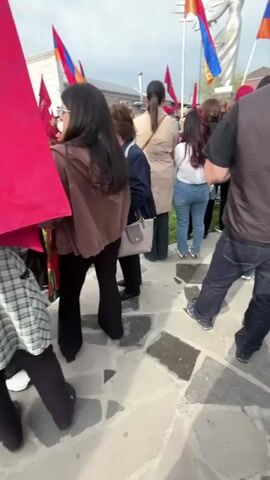 A crowd of people stands on a paved area near a large statue. Many individuals hold red flags with what appears to be text printed on them. Several Armenian flags are also visible. The statue is of a woman with outstretched arms and a radiant sun behind her. The weather appears overcast. The crowd is dense, and most individuals are facing the statue. The area around the statue is relatively clear, with buildings visible in the background. The statue is a focal point in the scene, and the flags seem to indicate a gathering of some sort, possibly a demonstration or commemoration.