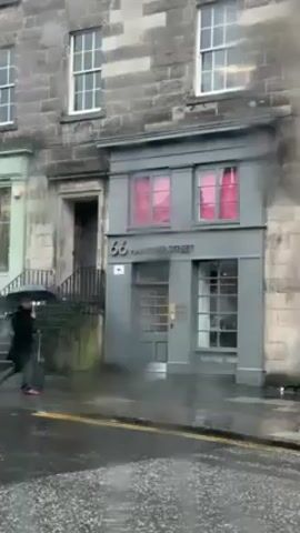 The video shows a street scene in the rain. A person with an umbrella and a rolling suitcase walks across the street in front of a building labeled "66 HAMILTON STREET". The building is gray, and the windows on the second floor have pink curtains. The street is wet, and there are raindrops visible on the camera lens. The building is made of stone, and there is another building to the left with green trim.