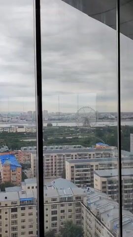 The video pans across a cityscape seen through a window. In the foreground, a vertical window frame divides the view. The cityscape is densely populated with buildings of varying heights and architectural styles. Some buildings are modern, while others appear older. Trees line the streets and are interspersed among the buildings, suggesting a city with significant green spaces. In the distance, a body of water is visible, and beyond that, more buildings extend to the horizon. A Ferris wheel can be seen on the right side of the distant skyline. The sky is overcast with clouds, indicating a possibly cloudy or gloomy day. The video captures the general ambiance of an urban environment. There are no people or specific actions clearly visible in the footage.