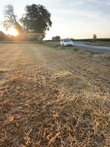 A car drives down a rural road next to a harvested field. The sun sets behind a tree on the left side of the frame. A woman in a pink top and denim shorts takes a selfie in front of a haystack. She smiles and poses for the camera, with the harvested field and sunset in the background.