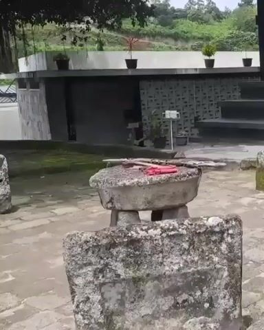 The video shows an outdoor scene with ancient stone structures and a modern building in the background. In the foreground, a large, round, flat-topped stone rests on three large, irregular stone supports, resembling a table. On top of this stone table are two wooden sticks and a folded red cloth. To the left and right of the stone table are other large, rough-hewn stones, some appearing to be upright slabs. The ground is paved with flagstones. In the background, a low-slung modern building with a flat roof and concrete walls is visible. The building has several potted plants on its roof. The overall environment appears to be an archaeological site or historical landmark.