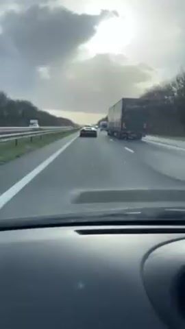 The view from inside a car driving on a multi-lane highway shows a dark-colored Lamborghini ahead in the same lane. A large truck is also in the lane to the right of the Lamborghini. The sky is overcast with the sun trying to break through the clouds. The highway appears to be bordered by trees and greenery.