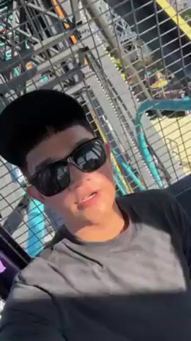 A young man with a black cap and sunglasses films himself on a Ferris wheel. He wears a gray t-shirt and has short dark hair. The Ferris wheel structure is visible around him, with metal bars and sections of seating in the background. Other rides are seen at a distance through the structure. The lighting is bright, indicating it's likely daytime.