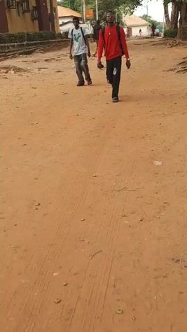 The video depicts a sandy, red-dirt road flanked by buildings and trees. Two men walk away from the camera at the beginning, followed by a group of three men further down the road. The road shows vehicle tire tracks and some scattered debris. The weather seems clear and bright.