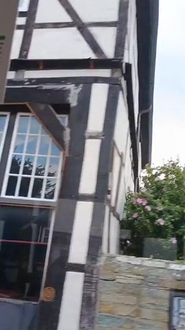 A video pans across a residential area with a mix of modern and historic buildings. The video begins with a view of a half-timbered house with white infill panels and dark timber framing, surrounded by lush green foliage and pink flowers. This transitions to a view of a modern, pale yellow building with a visible chimney and a brick wall in the foreground. The video then shows a closer view of the pale yellow building, highlighting its windows. Next, a different half-timbered building with white panels and dark timbers is shown, featuring a red door and a car passing by. Finally, the video pans to another half-timbered house with a distinctive gabled roof and dark timbers, set against a backdrop of trees and a stone wall. The sky is overcast throughout the video.