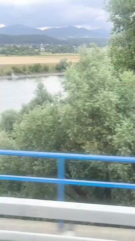 The video shows a view from a bridge overlooking a river and a landscape. The river is wide and has a gentle current. On the opposite bank, there are fields, trees, and a town with buildings and a church spire. In the distance, rolling hills or mountains are visible under a cloudy sky. The foreground of the video shows the blue railing of the bridge and some greenery. The camera appears to be moving, suggesting the view is from a vehicle or train.