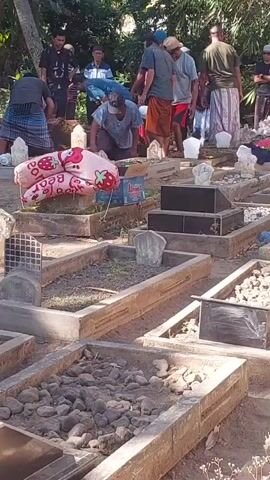 In an outdoor cemetery setting, a group of men are gathered around a grave. Some of the men are wearing traditional Islamic clothing. One man is kneeling at the grave, appearing to be tending to it. Another man is standing nearby, also leaning towards the grave. A pink balloon with a strawberry design and the word "BEAR" is placed on the grave. Several other graves are visible in the foreground and background, some adorned with stones or small white markers. The environment is filled with greenery, suggesting it is a natural outdoor space.