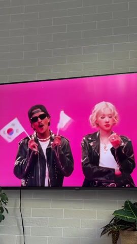 A screen displays a music video with a pink backdrop. In the foreground, a person wearing a black leather jacket, sunglasses, and a backwards baseball cap holds a microphone and a white flag with a face on it. To their right, a person with blonde hair, also wearing a black leather jacket, plays a drum set. The screen is placed on a white brick ledge, with green plants visible at the bottom.