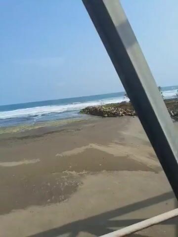 The video shows a view from a moving vehicle, possibly a train, as it passes by a beach and the ocean. The beach appears sandy with some rocks and sparse vegetation. In the distance, waves are breaking on the shore, and the sky is clear and blue. The vehicle's structure, such as window frames or support beams, are visible in the foreground, partially obstructing the view of the scenery. The camera angle is slightly downward, looking out towards the beach and sea. There are no people visible in the video. The overall impression is of a coastal landscape on a sunny day, viewed from a mode of transportation.