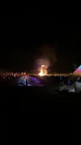 A large bonfire is burning brightly in the center of the frame, with flames reaching high into the dark night sky. Sparks and smoke billow upwards. A crowd of people is gathered around the bonfire, silhouetted against the light. In the foreground, a striped tent with purple lights draped over it is visible. The ground appears to be uneven and dark. The overall impression is of a nighttime outdoor gathering or festival centered around a large fire.