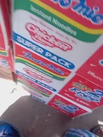The video shows a roadside stand selling Indomie instant noodles. Boxes of chicken-flavored Indomie are visible. Bundles of Indomie packets are arranged on a wooden shelf. Street noise can be heard. The camera moves quickly.