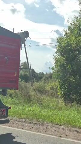 A tractor is seen from behind as it drives on a dirt road through a rural area. The tractor is red and appears to be pulling a trailer. The surrounding environment is green with trees and grass. The sky is cloudy with patches of blue. A power pole and wires are visible in the background. The video is shot from a moving vehicle, likely following the tractor.