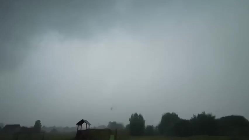 This media file shows a rural landscape in Kyviškės, Lithuania. The scene is dominated by a dark, overcast sky, suggesting inclement weather, possibly a thunderstorm approaching or in progress. In the foreground, there is a small wooden playhouse with a roof and open sides, and part of a fence is visible on the left. Further in the background, several trees create a silhouette against the hazy sky, and a few indistinct structures can be seen. The overall atmosphere is somber and muted due to the heavy cloud cover and the low visibility, which blurs the details of the landscape. There is no visible text in the media file.