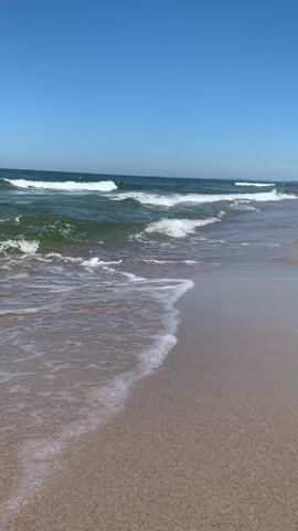 This is a vertical video of a beach scene. The subjects are the ocean and the sand. The scene is a beach with the ocean in the foreground and the sky in the background. The setting is a beach, likely in Baltiysk, Russia, given the provided context.

The activity taking place is the gentle lapping of waves on the sandy shore. The water is a deep green with white foam as the waves break. The sky is a clear, bright blue, suggesting a sunny day. The time of day appears to be daytime, likely mid-morning to afternoon, due to the clear blue sky and lack of shadows. The weather seems to be warm and pleasant.

Notable details include the texture of the sand, which is a light brown and appears somewhat wet near the water's edge. The waves are not large, indicating calm conditions. There are no visible objects or people in the immediate foreground, but in the distance, there are a few small figures that appear to be people in the water, possibly swimming or wading. The overall emotion conveyed is one of tranquility and relaxation. There is no visible text in the media file.