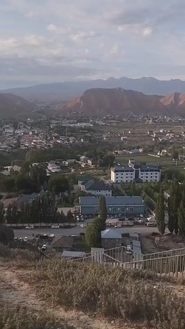 A panoramic video captures a wide view of a valley town nestled between arid mountains under a clear, partly cloudy sky. The camera pans from right to left, revealing a tiered landscape. In the foreground, a steep incline covered in dry brush and plants leads down to a set of metal stairs with a railing. Several individuals, dressed in casual clothing, are visible on these stairs and a viewing platform at the bottom.

As the camera continues to pan, the town unfolds below. Buildings of various sizes and styles, including modern apartment blocks and smaller houses with colorful roofs, are scattered throughout. Roads with vehicles, including cars and what appear to be trucks, wind through the urban area. Greenery, such as trees and fields, is interspersed with the buildings, suggesting a mix of residential and possibly agricultural zones.

The middle ground features more of the town and a river or waterway flowing through it. A bridge can be seen crossing this water. Tall, slender trees line some of the streets and green spaces.

In the background, imposing mountains dominate the horizon. Their slopes are a reddish-brown, indicating an arid climate, and some of the higher peaks are capped with snow, contrasting sharply with the desert-like terrain. The sky above is a bright blue with scattered white clouds, and the lighting suggests it is either early morning or late afternoon, with soft sunlight casting a warm glow over the scene. The overall impression is one of a tranquil, somewhat remote town situated in a striking natural landscape.