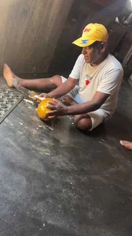 A man kneels on a dark floor, a yellow coconut in his hands. He wears a white t-shirt and blue shorts, with a yellow baseball cap. He uses a large knife to cut the coconut, which is yellow and appears to be peeled in places. Several pieces of the coconut peel are on the floor around him. The background is dimly lit, with wooden walls.