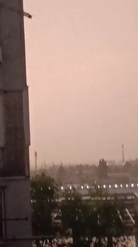 The media file shows a city skyline at dusk or dawn, with a hazy, light pink sky. In the foreground, a portion of a brick building is visible on the left side, with a railing or fence in front of it. Beyond the building, the city stretches out with buildings and trees. Lights are visible in the distance, suggesting urban activity. Tall structures, possibly communication towers or cranes, punctuate the skyline. The overall impression is one of a slightly overcast or dusty atmosphere. The video abruptly cuts to black. No people are visible in the frames.