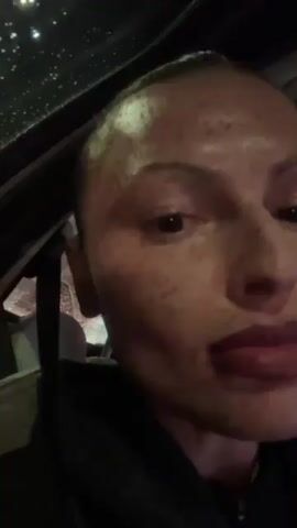 A woman is inside a car, looking directly at the camera. The car's ceiling has a sunroof, through which one can see rain or water droplets and some blurry lights. The woman has her hair pulled back, and she has freckles on her face. She makes a kissing face and then smiles. She is wearing a black garment.