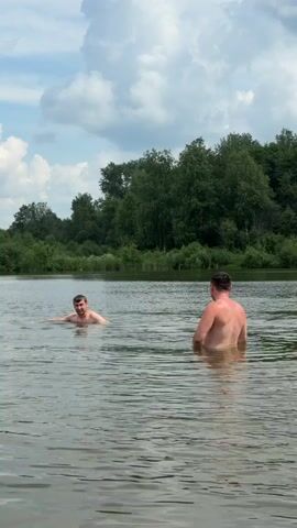 Two men are swimming in a lake. The man on the left is closer to the camera and is facing it, swimming towards the camera. The man on the right is further away from the camera and is facing away from it, standing in the water up to his waist. The lake is calm and the water is clear. The background is a line of trees under a cloudy sky. The video is shot from a low angle, close to the water's surface.