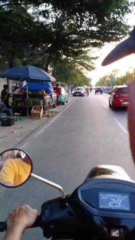 A first-person perspective video shot from a motorcycle shows a street scene at dusk. The motorcycle is traveling on the right side of a two-lane road. A line of parked cars is visible on the left side of the road, along with a sidewalk and trees. Several people are seen on the sidewalk and near the parked cars. The motorcycle's digital speedometer displays a speed of 28, then 26, then 25 kilometers per hour. In the mirror on the left side of the motorcycle, a person wearing a yellow shirt is visible. The sky is visible above the trees, indicating it is either sunrise or sunset.